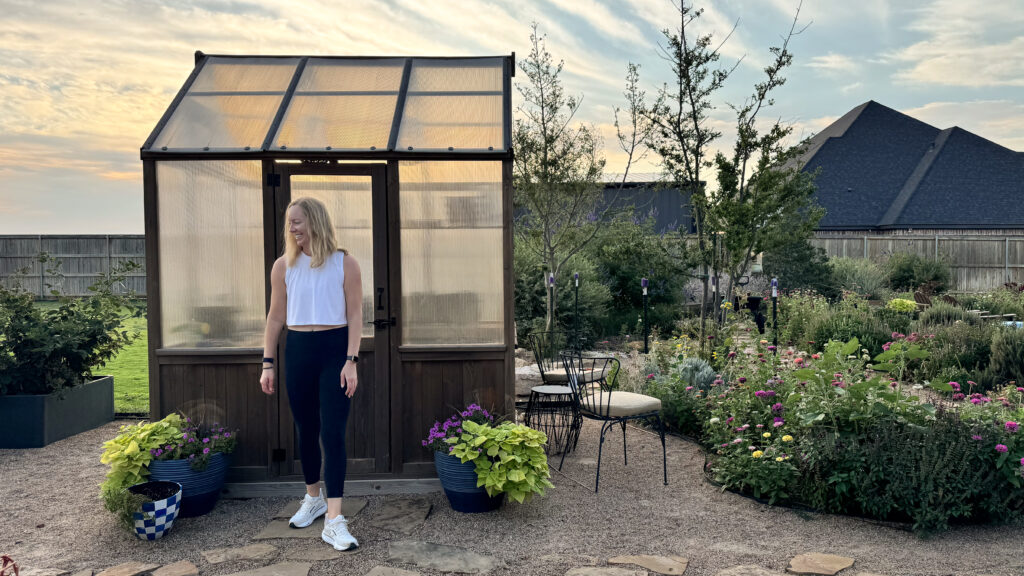 Julia standing in the garden in front of a green house with morning light. To the right, flower beds and trees fill the frame